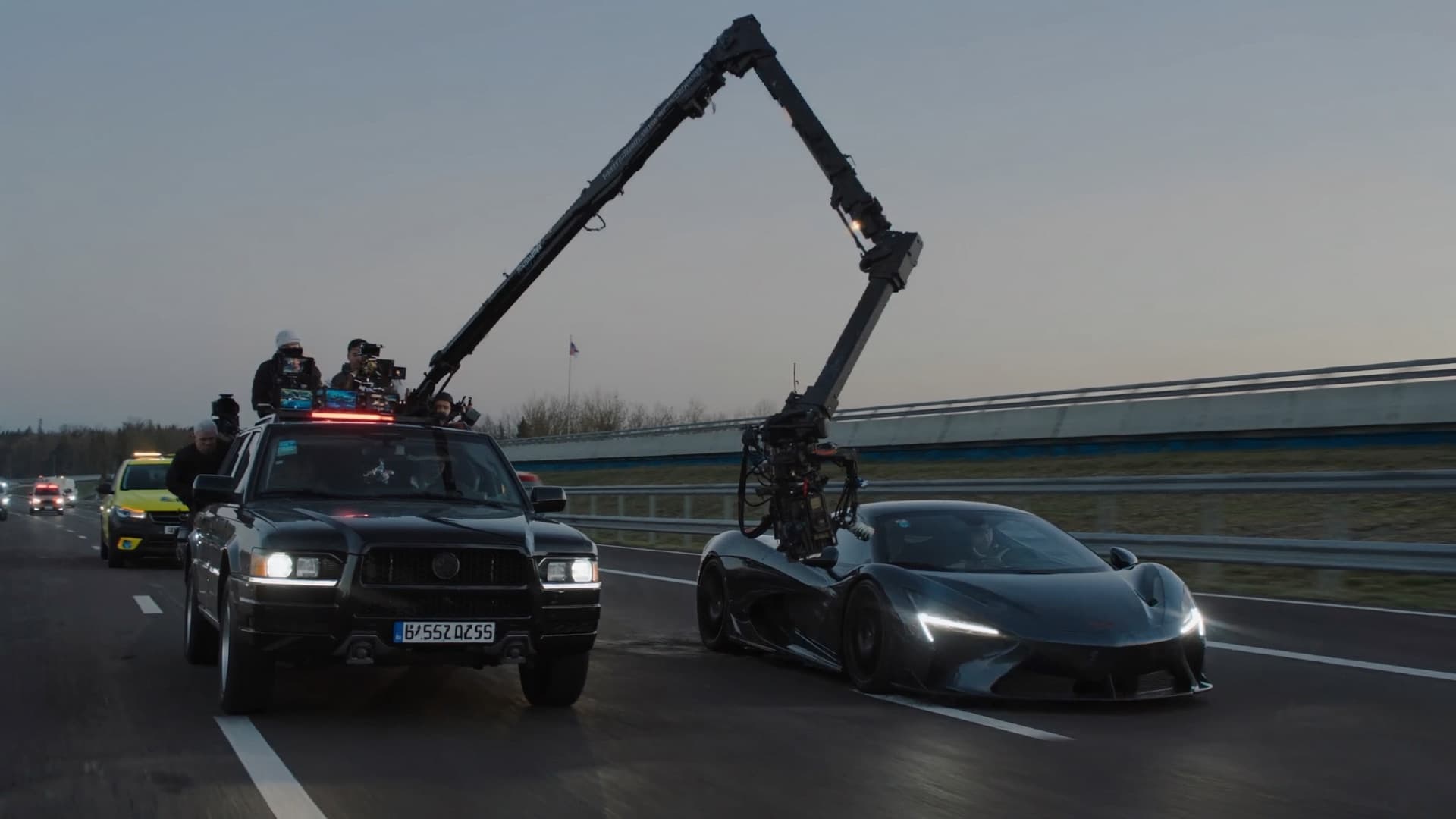 A camera crew on a tracking vehicle filming a car on a closed highway at dusk