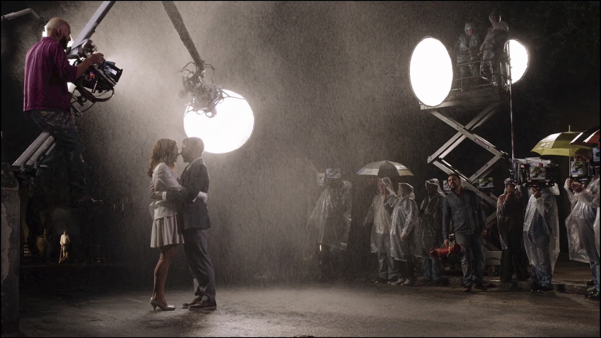 A night exterior on set with rain towers and lights over two actors