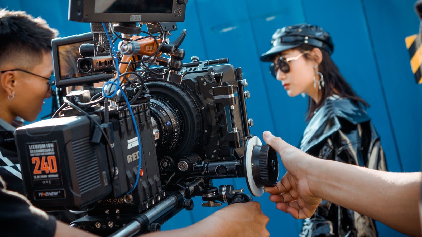 A camera assistant pulling focus on a cinema camera while an actor waits in front of a blue backdrop